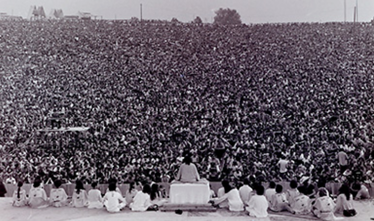 Uma fotografia mostra a vista traseira de um palco, em frente ao qual uma grande multidão está reunida. Swami Satchidananda está sentado de pernas cruzadas no topo de um pódio, com um pequeno grupo de outras pessoas sentadas no palco a cada lado dele.