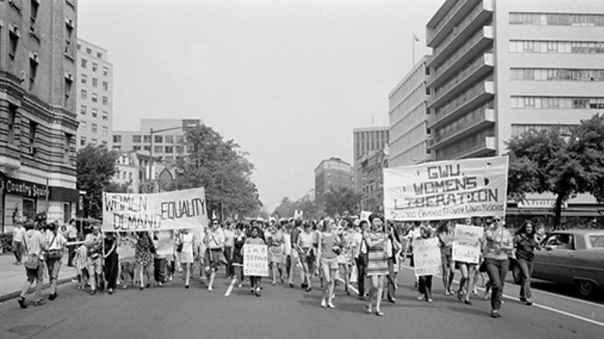 Uma fotografia mostra uma marcha de protesto de mulheres em uma rua da cidade. Os participantes seguram cartazes com mensagens como “Mulheres exigem igualdade”; “Sou cidadã de segunda classe” e “Libertação das Mulheres da GWU. Estudantes, funcionários, professores, esposas, vizinhos.