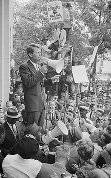 Uma fotografia mostra Robert Kennedy falando para uma grande multidão por meio de um megafone.