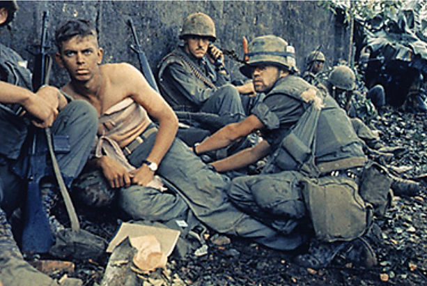 Uma fotografia mostra um grupo de soldados americanos uniformizados agachados ao lado de uma parede. Um soldado está sem camisa, com uma grande bandagem enrolada no peito.