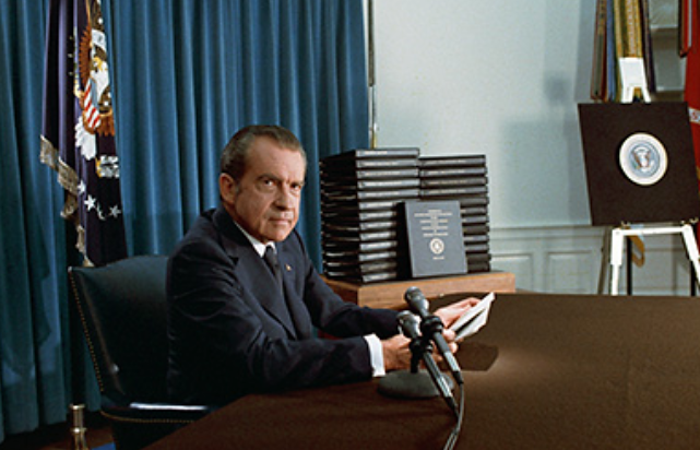 Uma fotografia mostra o presidente Nixon sentado em uma mesa ao lado de vários microfones, segurando papéis enquanto se prepara para se dirigir à nação.