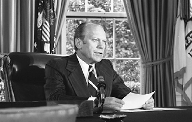 Uma fotografia mostra Gerald Ford sentado em uma mesa com uma folha de papel à sua frente, falando ao microfone.