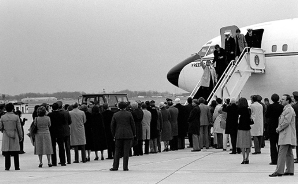 Uma fotografia mostra ex-reféns descendo um lance de escadas para sair de um avião oficial; uma multidão de pessoas os espera no chão.