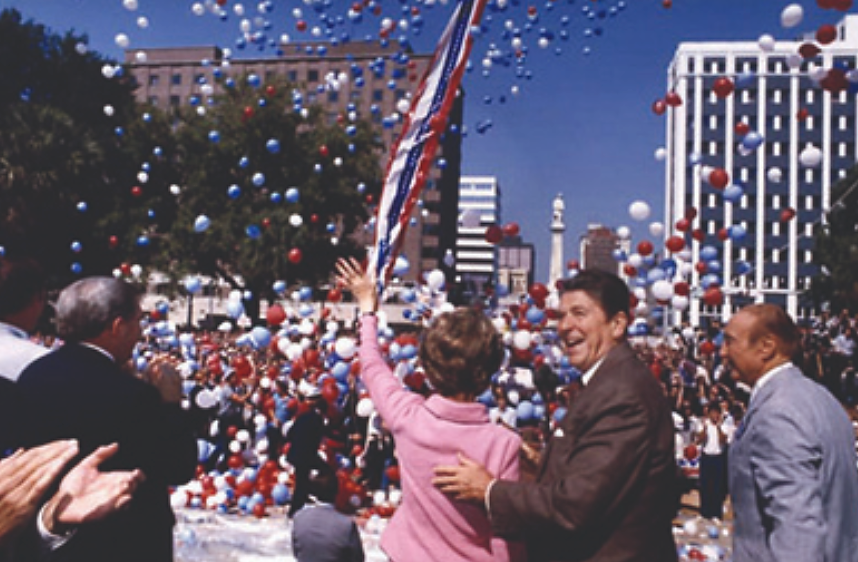 一张照片显示罗纳德和南希·里根在竞选活动中。 他们站在欢呼的人群中，周围环绕着红色、白色和蓝色的气球。 南希·里根向人群挥手致意；罗纳德·里根微笑着把手放在她的背上。