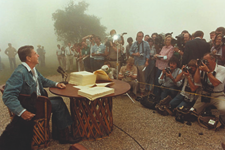 A photograph shows Ronald Reagan signing legislation while seated outdoors at a rustic table. He is dressed in blue jeans, a denim jacket, and cowboy boots, and he strokes the head of a large black dog seated beside him. In front of Reagan, the press takes photographs.