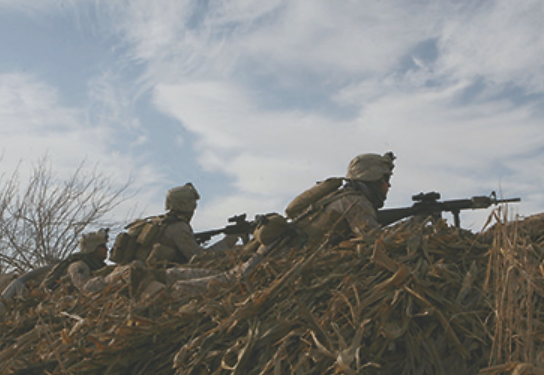 Uma fotografia mostra vários fuzileiros navais posicionados atrás de uma colina, com suas armas prontas para o ataque.