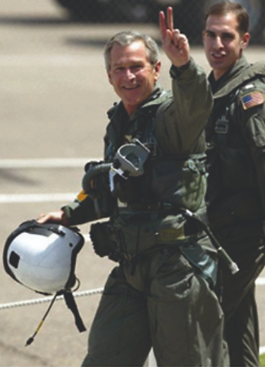 A photograph shows George W. Bush walking with naval flight officer Lt. Ryan Phillips following the president’s arrival on the USS Abraham Lincoln. Bush wears a flight suit and gives a victory symbol to the cameras with his hand.