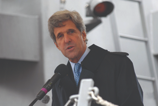 A photograph of John Kerry speaking into a microphone is shown.