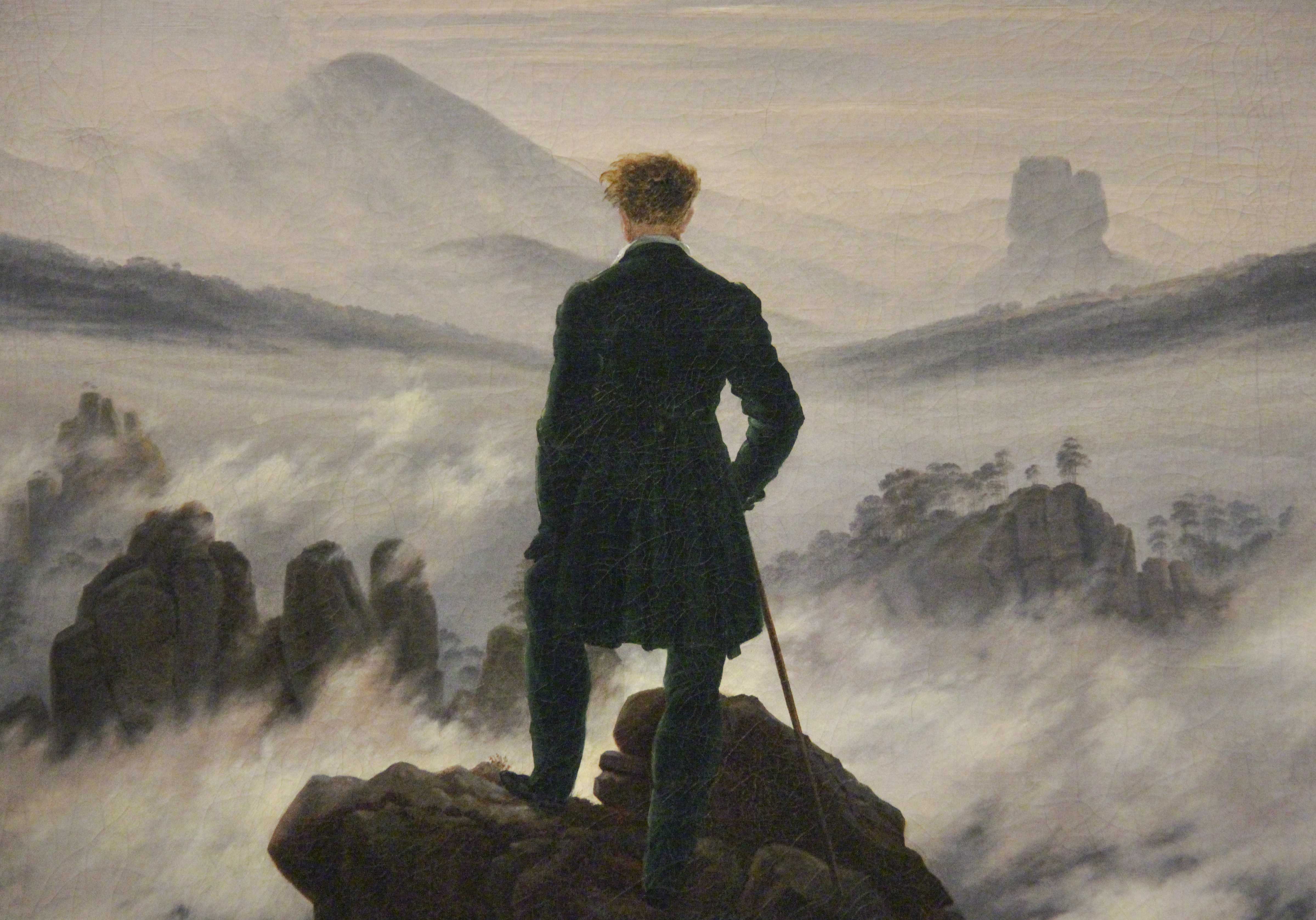 man in dark suit stands on mountain overlooking a sea of clouds
