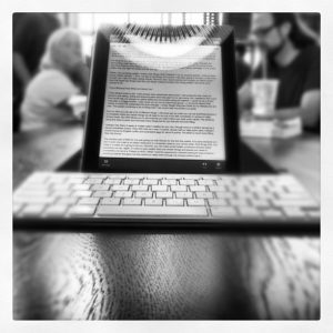 Black and white photo of a tablet displaying text and wireless keyboard, on a table in a cafe with people in the background.
