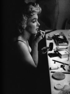 Black and white photo of Marilyn Monroe applying makeup in a dressing room.