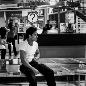 A young man sitting underneath a question mark sign, positioned as if he is posing a question himself.