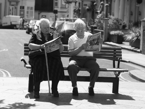 Black and white photograph of two people sitting on a bench near a street, reading newspapers