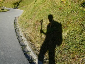Photo showing a shadow of a man carrying a walking stick. The shadow appears on a grassy hill alongside a road.