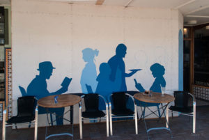 Blue silhouettes of people sitting, walking, and reading painted on a wall behind a common area with tables and chairs
