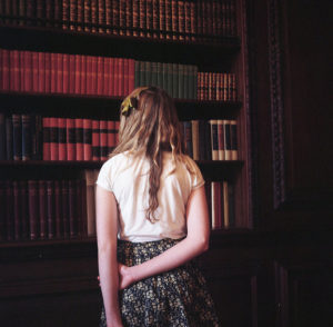 Blond woman standing with her back to the camera, facing a tall bookshelf lined with leather-bound volume sets