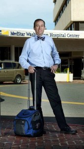 Photo of a man in a bow tie, dress shirt, and slacks standing outside the front of MedStar Washington Hospital Center, with his hand on the extended handle of a wheelie bag.