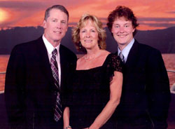 Family photo: father, mother, and teenaged son from left to right. The men wear suits and the woman wears a black dress. They are standing, posed against a sunset backdrop.