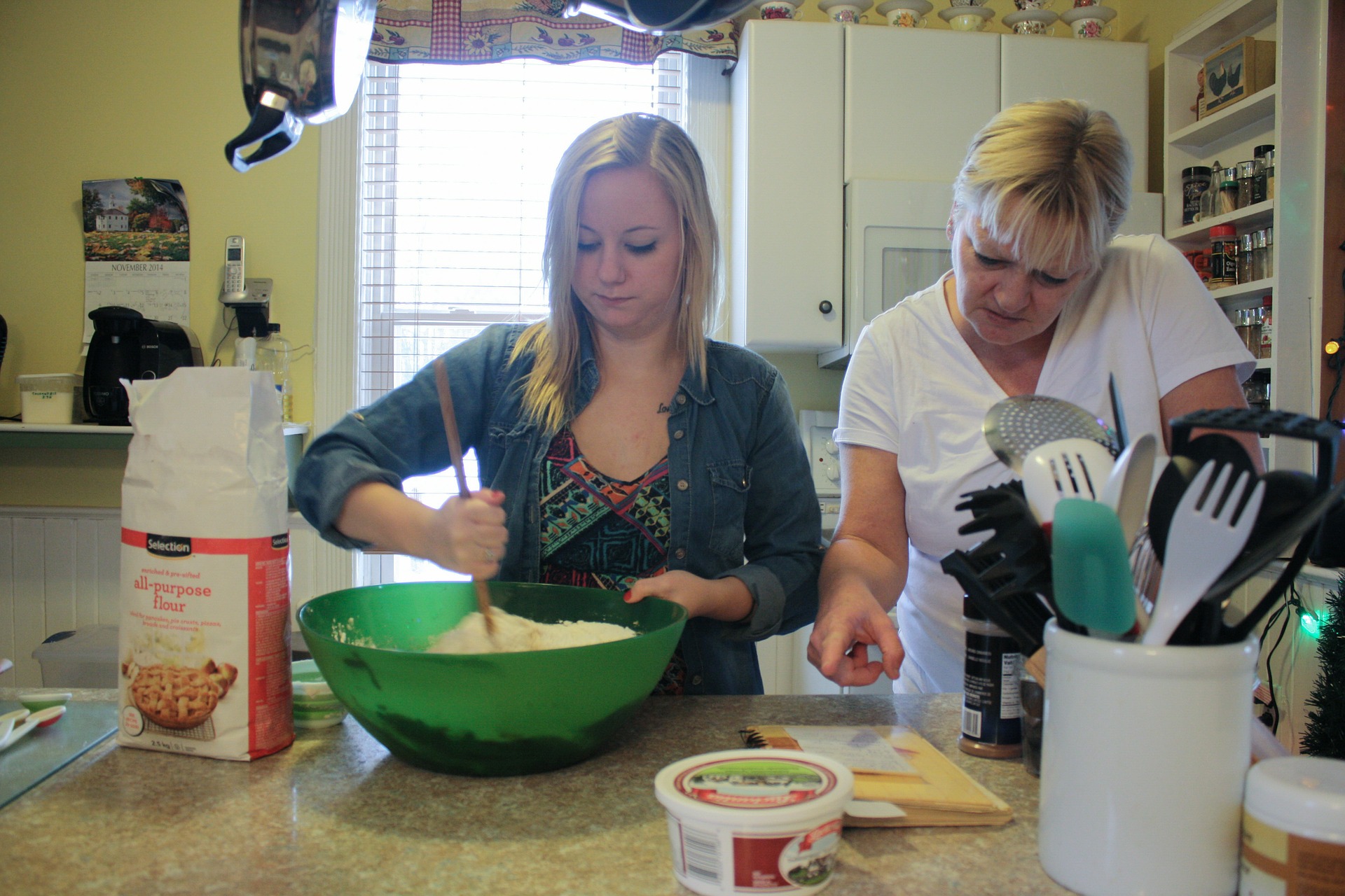 a girl string ingredients in a mixing bowl while a woman looks at a recipe