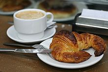 Coffee in a mug next to a croissant on a plate for breakfast.