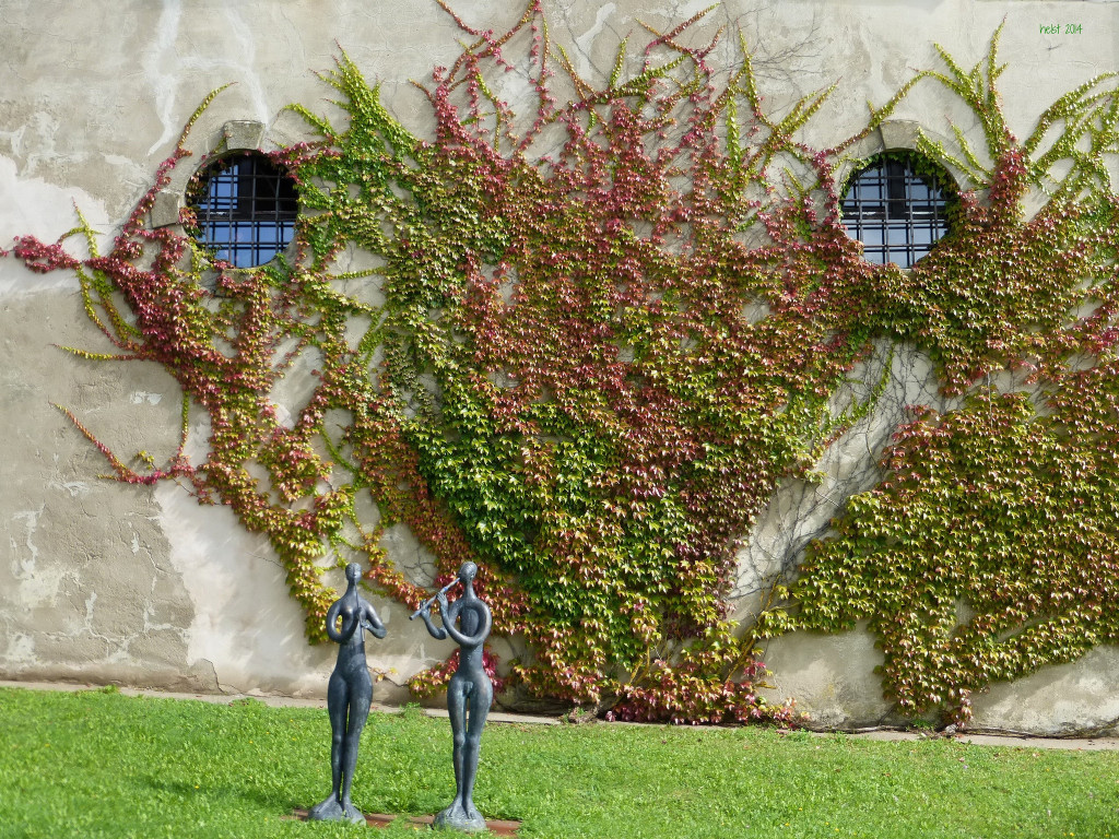 Photo of a vine-covered wall of Burg Spielberg; on the lawn in the foreground are two small bronze sculptures of a nude man and woman; the man is playing a flute.