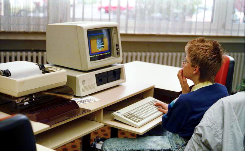 A boy sitting in front of a personal computer; the computer is considerably blockier than computers today.