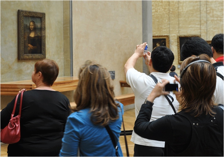 Image depicts several people standing and looking at museum displays. La Joconde (aka “The Mona Lisa”) appears on a wall in the background.