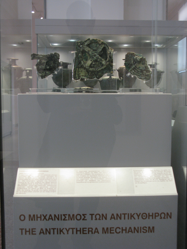 Photo of The Antikythera Mechanism exhibit at the National Archeological Museum of Athens. Grecian urns are on display in the background.