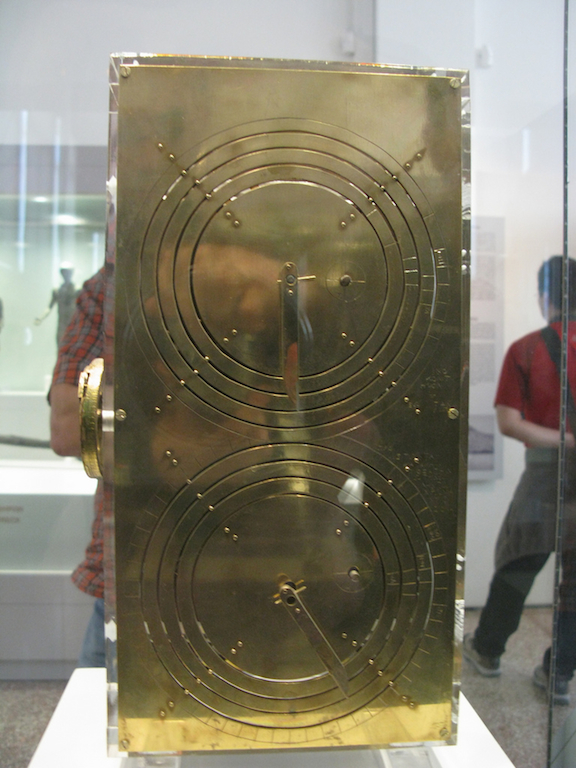 Photo of a display case containing a metallic object with two sets of concentric circles inscribed on its face, and dials akin to those of an analog clock or watch. Museums patrons in background.
