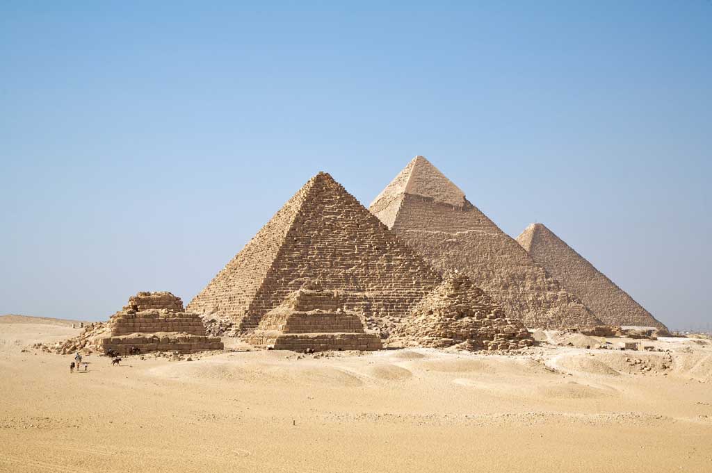The picture is that of the six Pyramids of Giza. In the background are the three large pyramids of kings Menkaure, Khafre, and Khufu situated in a line. All three are smooth-sided. In front, are three smaller pyramids, of which only one is not a stepped pyramid.