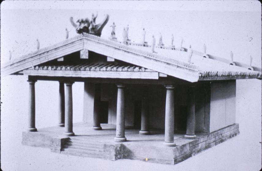 Picture of a model Etruscan temple. The temple has a slanted roof stationed atop eight columns and an inner chamber. The temple itself sits atop a porch with staircase.