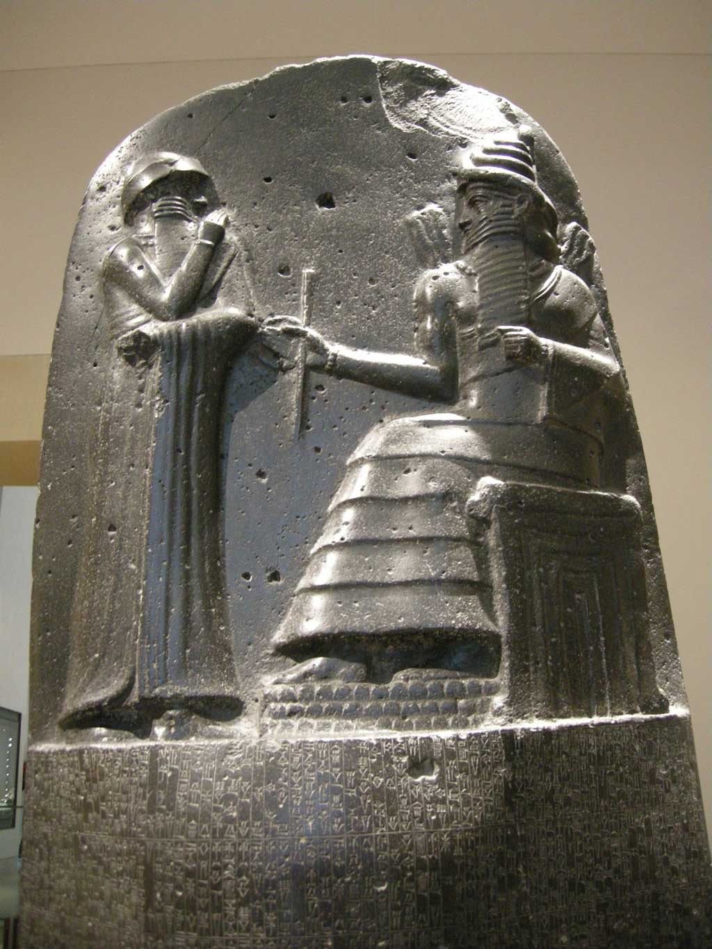 Picture of Hammurabi's Code. A dark cylindrical stone statue with a carving on the upper portion of the Code. Here, one sees a representation of king Hammurabi receiving his law code from the god Shamash who sits before him. Both are two-dimensional in form.