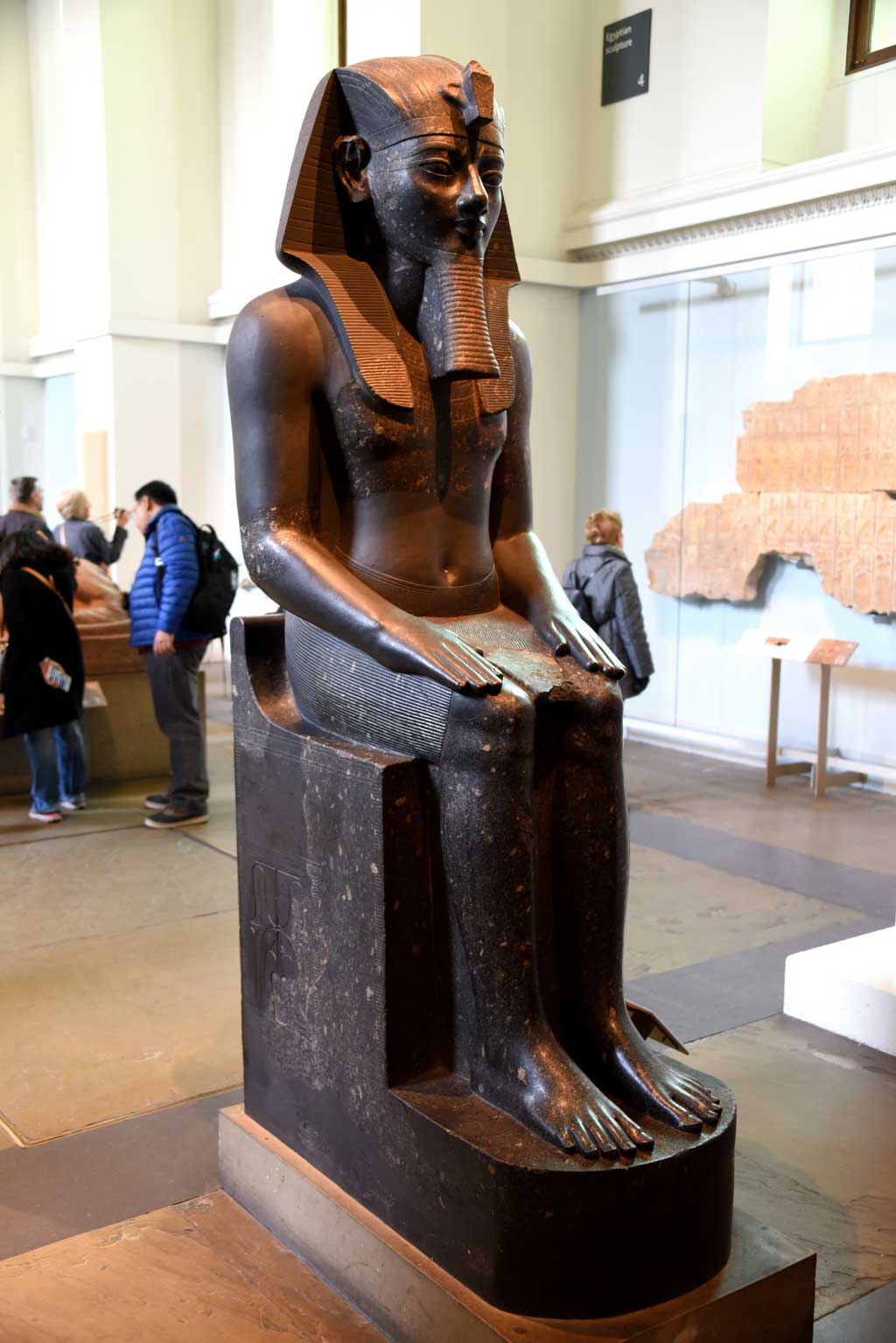 The picture is of a seated Amenhotep III, carved out of black stone. Here, the king sits while looking straight ahead, with hands placed flatly on his bent knees.