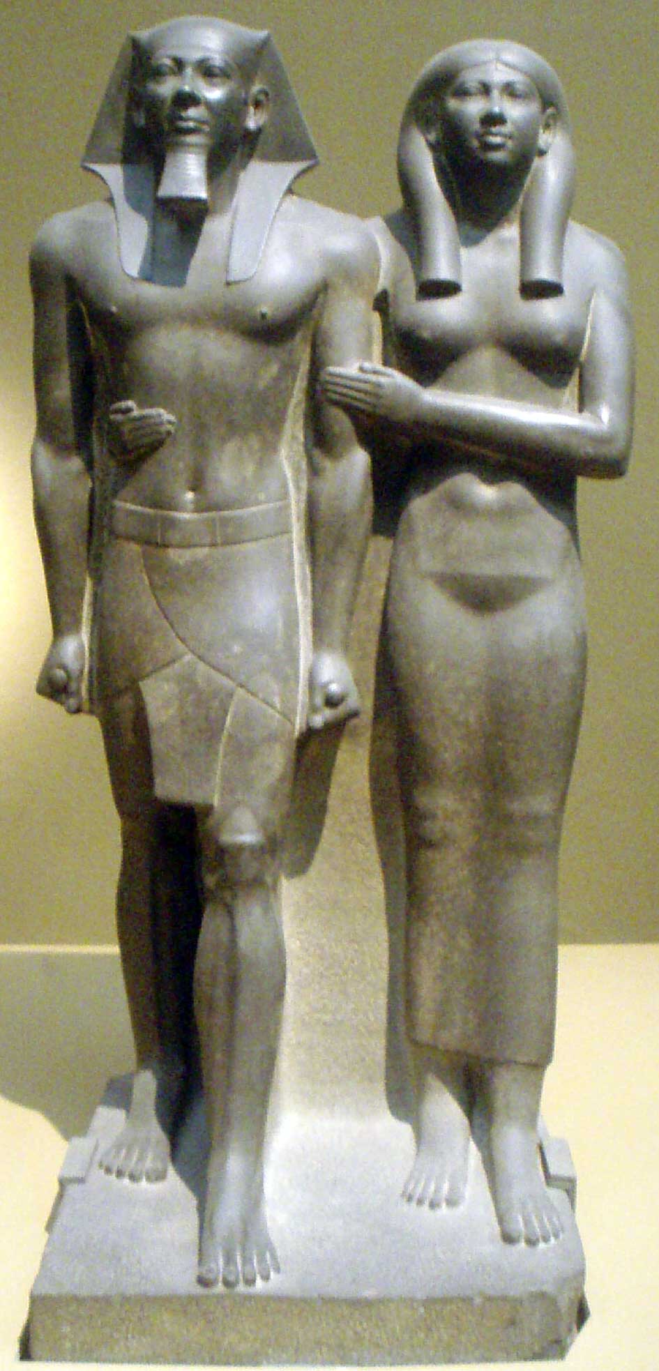 The image is that of the Egyptian king Menkaure and his queen, Khamerernebty II. Hewn from dark stone, both king and queen are stoic in appearance and possess ideal bodies. Although standing shoulder to shoulder, Menkaure's foot steps ahead of his wife to illustrate his superior position.