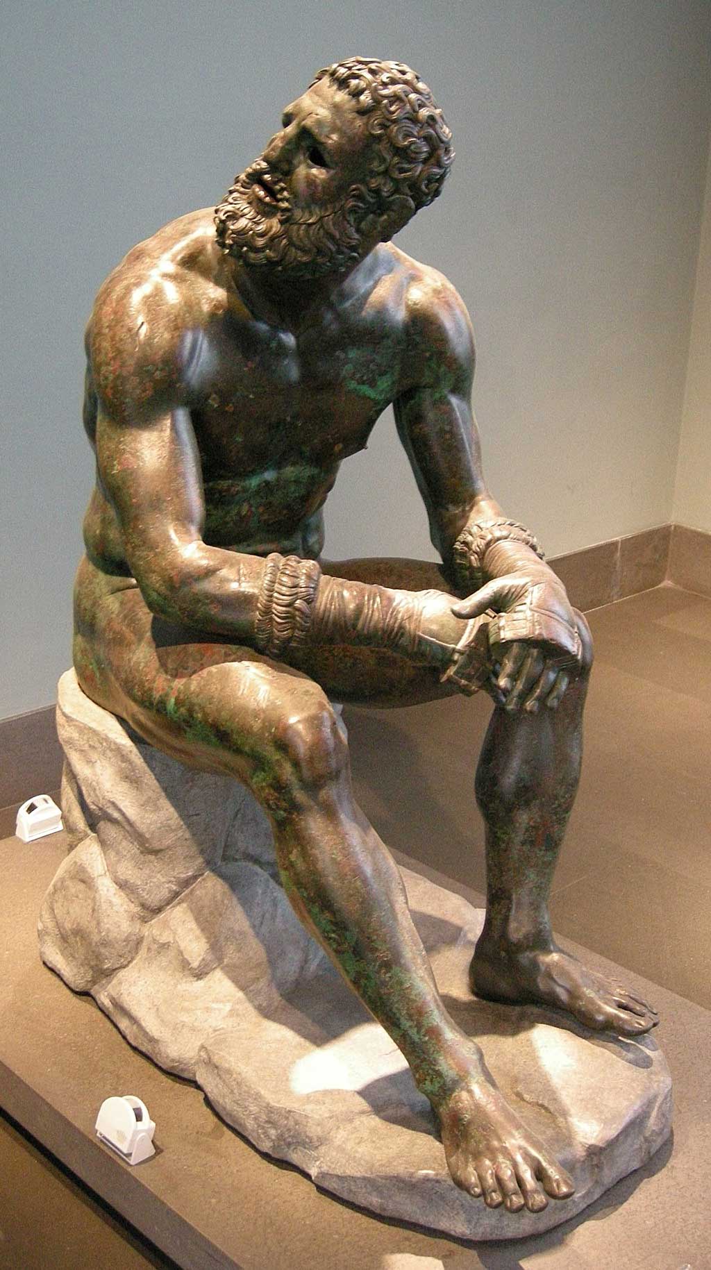 Image of the Boxer statue. The bearded man sits on a stone, looking upwards while wearing the leather gloves still strapped around both hands.