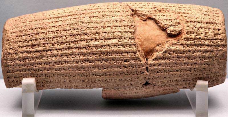 Image of the Cyrus Cylinder. The tiny cylinder is in the shape of a stone tube sitting atop a platform in a museum.