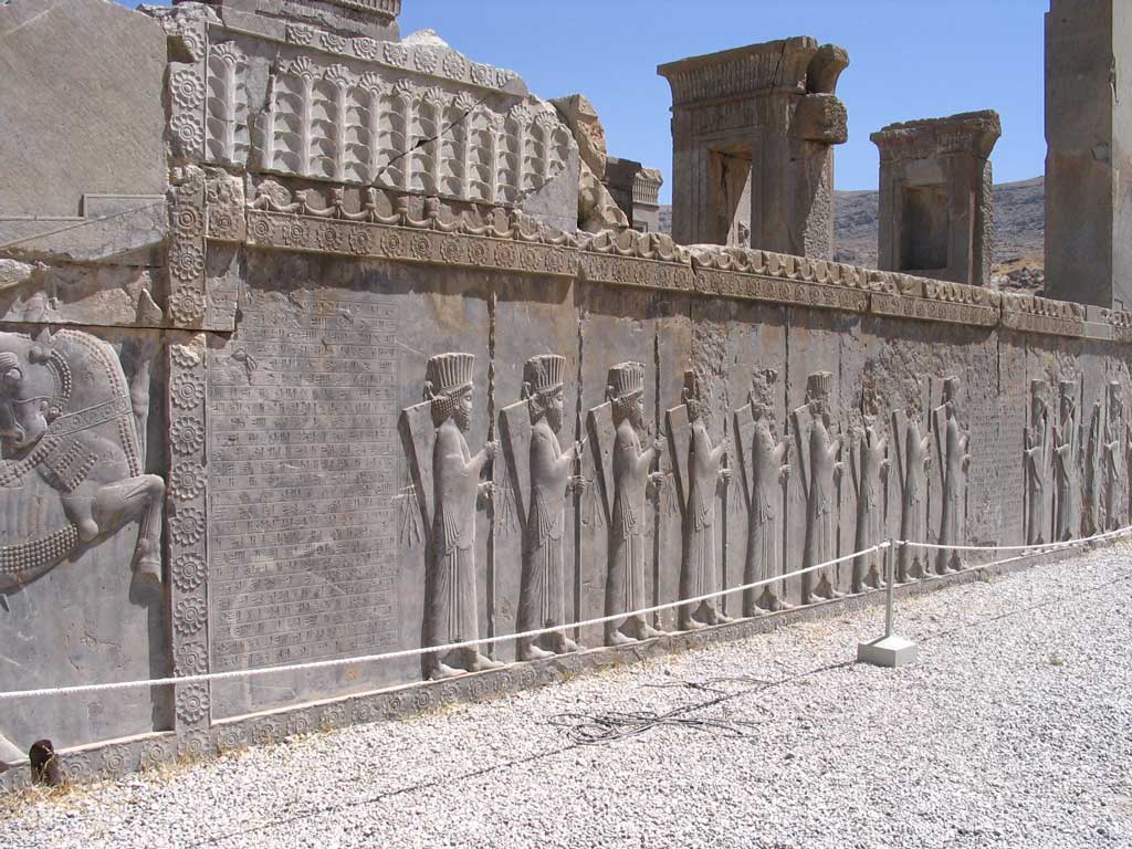 Image of grey stone wall relief depicting two lines of two-dimensional Persian officials facing one another. The remains represent one of the few from the Persian capital of Persepolis.