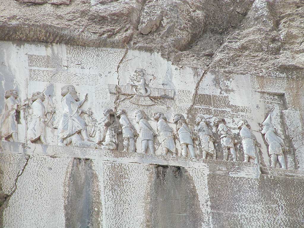 Image of the Behistun Inscription. Here, a two-dimensional Darius stands taller than the other figures in the scene as he recounts his ancestry and triumphs. The speech can be seen above their heads, written in three different cuneiform scripts.
