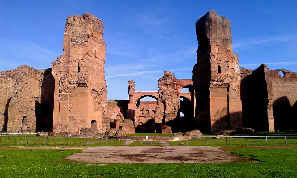 Picture of the outer remains of the Baths of Caracalla. In the foreground, there is an open courtyard leading up towards the entrance to baths. In the background are the remains of the baths themselves, with two large towers standing at the entryway of the principle facade and the great courtyard just beyond it.