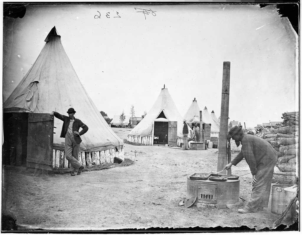Civil War soldiers preparing a meal in camp. Cooks work on makeshift stoves while other look on from tent barracks.