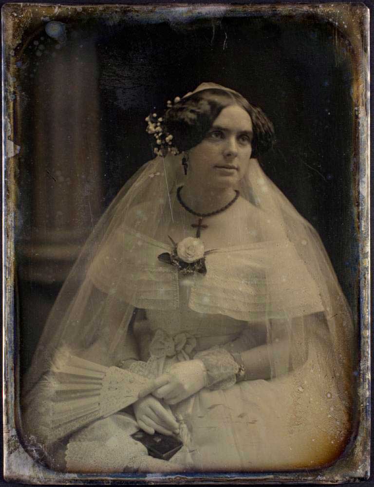 Old photo of a bride wearing a wedding dress and veil, holding a fan