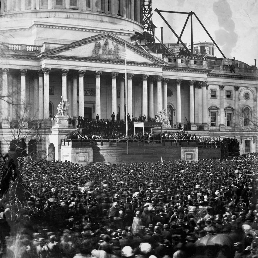 Photograph shows participants and crowd at the first inauguration of President Abraham Lincoln, at the U.S. Capitol, Washington, D.C. Lincoln is standing under the wood canopy, at the front, midway between the left and center posts. His face is in shadow but the white shirt front is visible.