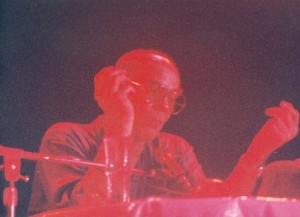 Low-quality photo showing Thompson seated at a table in front of a microphone. His figure appears in red tones, and the background is purple.