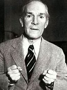 Black and white photo of Upton Sinclair, in a suit, looking straight into the camera. Both his hands are formed as fists, as if he's sincerely trying to explain something