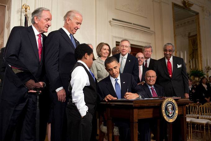 Several men and women standing around President Obama signing the Affordable Care Act sitting at a desk.