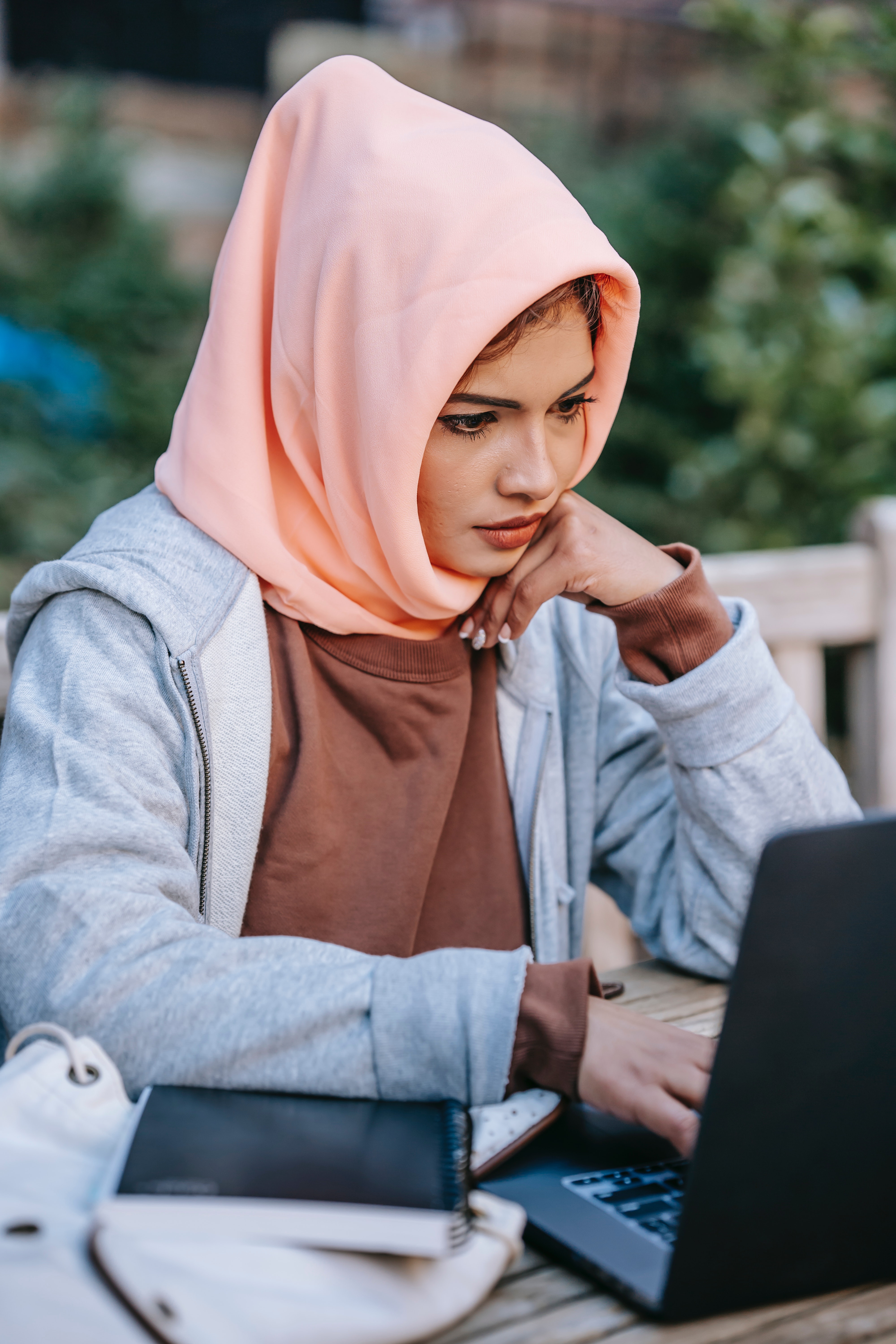 Una joven seria con un pañuelo en la cabeza, mirando la pantalla de una computadora y escribiendo en una mesa al aire libre.