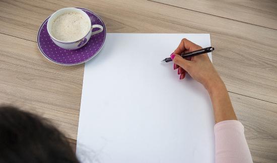 A hand grasps a pen over a blank sheet of paper with a cup of coffee next to it.