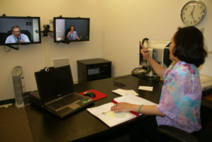 A man and a woman teleconferencing. The woman is sitting at a desk. There is a clock on the wall.