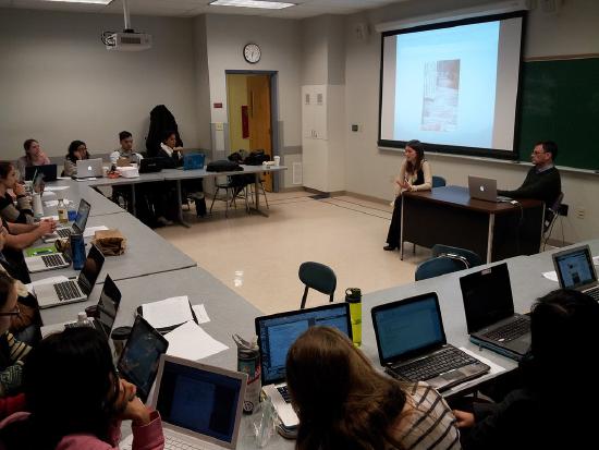 Photo of a classroom. Students are sitting at tables around the room with laptops in front of them. The teacher is at the front of the room, and an image is projected on a screen in front of the chalkboard. There are no windows, but there is a door and a clock.