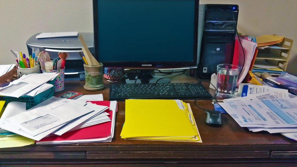 A fairly cluttered desk with a computer, folders, papers, pens, and pencils